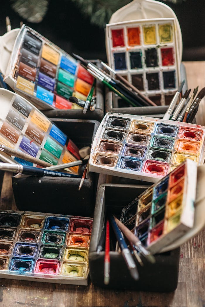 A collection of colorful watercolor paint sets and brushes arranged on a wooden table.