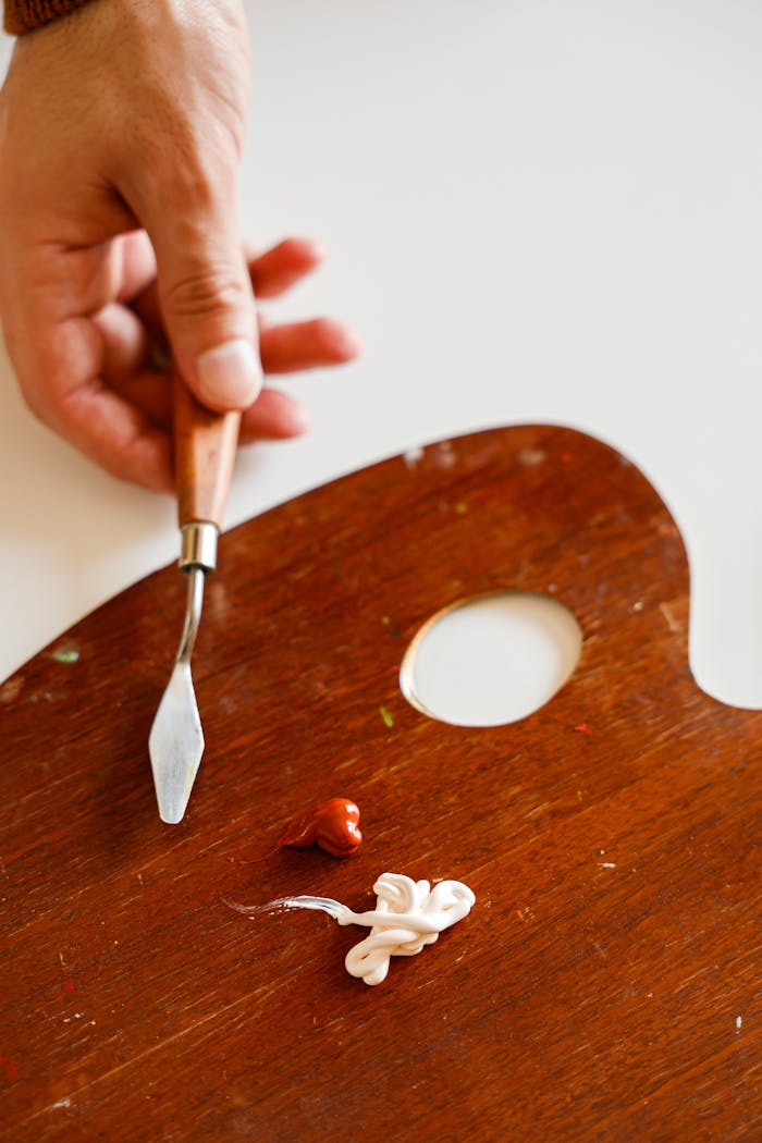 An artist's hand holding a palette knife with paints on a wooden palette for painting creativity.